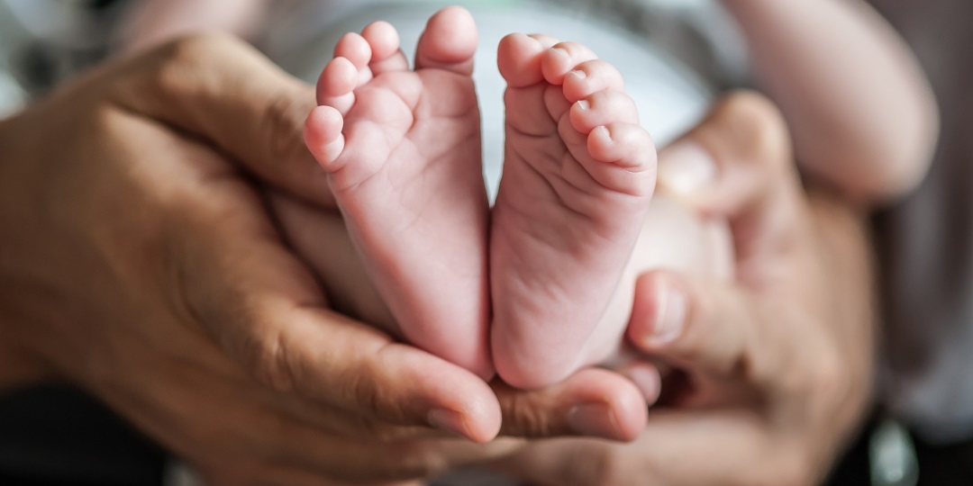 Mãos adultas seguram bebê, com seus pés em primeiro plano