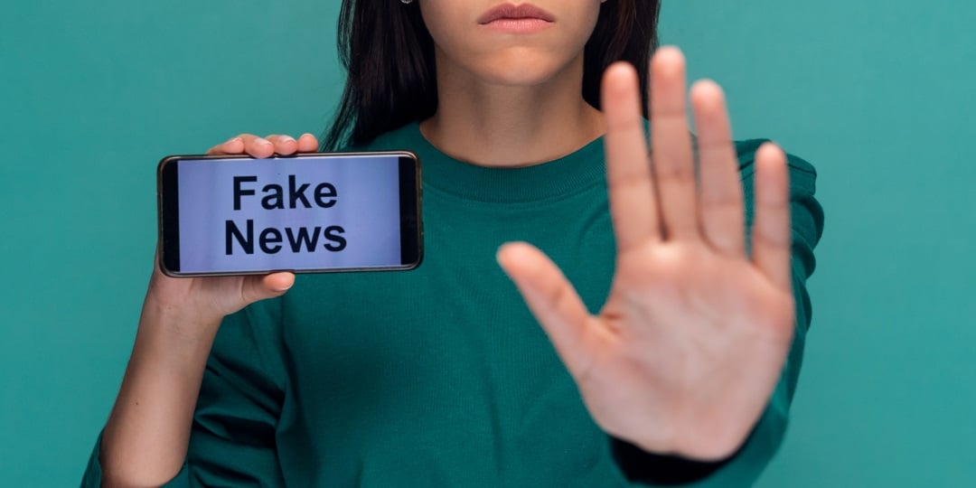 Mulher faz sinal de pare com uma mão e segura celular com "fake news" escrito na outra