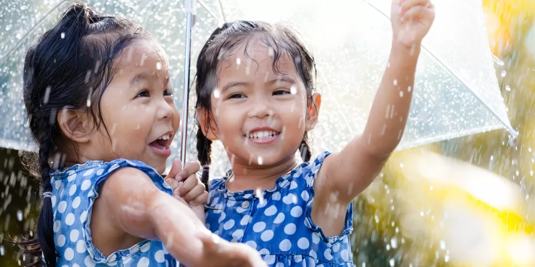 Duas crianças segurando um guarda-chuva e brincando com as gotas de chuva